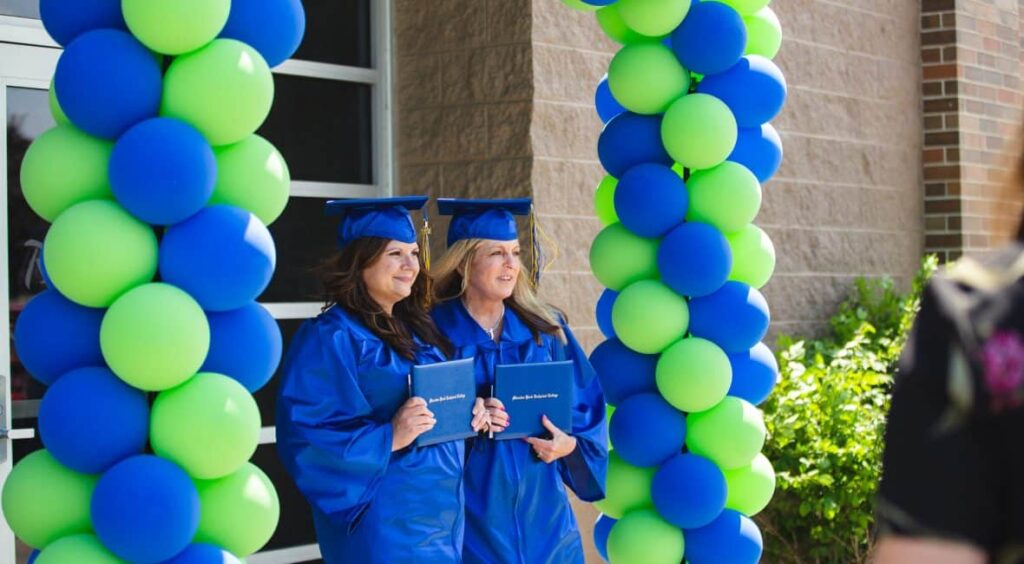 Two students in graduation gowns standing under a blue and green balloon arch presenting their degree documents. 