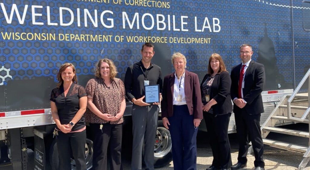 Group photo of DOC and Moraine Park employees in front of wall saying "Welding Mobile Lab."