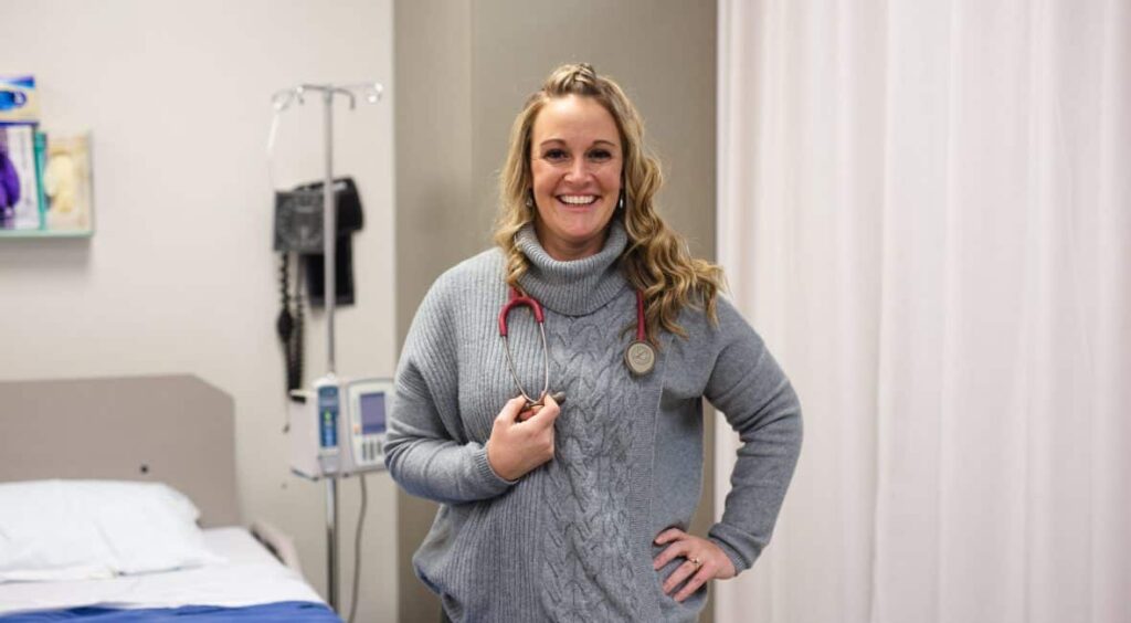 Kelly Hagner, Nursing student, standing in front of a hospital bed and smiling into the camera with a stetoscope around her neck.