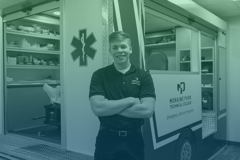 Male student crosses his arms while standing in front of a simulation ambulance and smiles at the camera