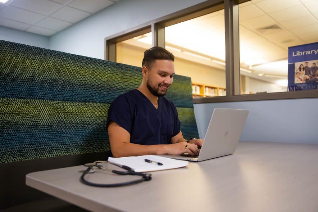 Male student sitting in a booth with a laptop. He is smiling and studying.