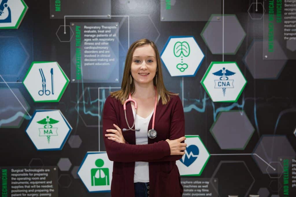 Female students standing in front of wall that shows symbols of different health science areas. She is crossing her arms and confidently looks into the camera.