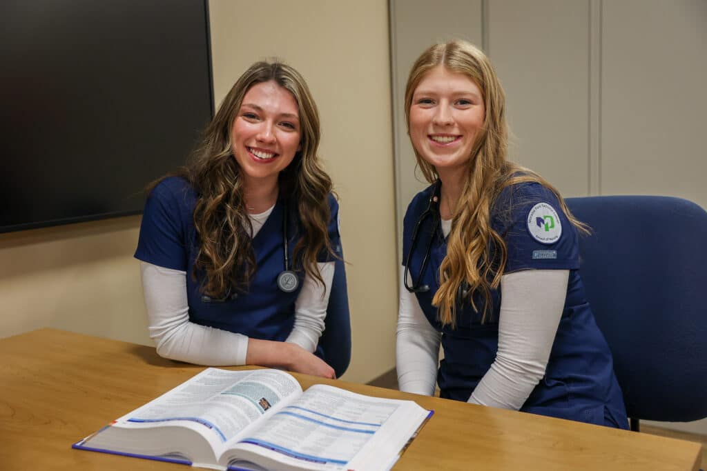 Two nursing students sitting at a table with an open book. They are studying for an exam and smiling into the camera.