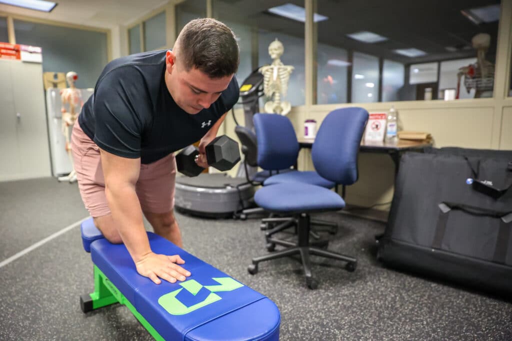 Health and Wellness student working out with a dumb bell bell while kneeing on a workout bench with one leg.