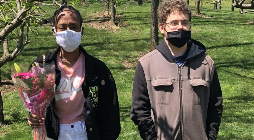 Two students standing outside in the green. One is holding flowers.