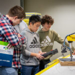 Three students playing with a joystick controlling a robotic arm.
