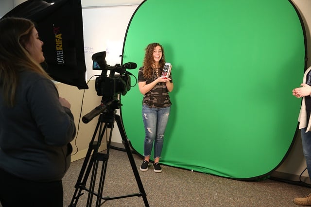 Marketing students standing in front of green screen and presenting a product. The video camera is pointing at her. 