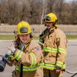 Two students in full firefighter gear carrying a hose.