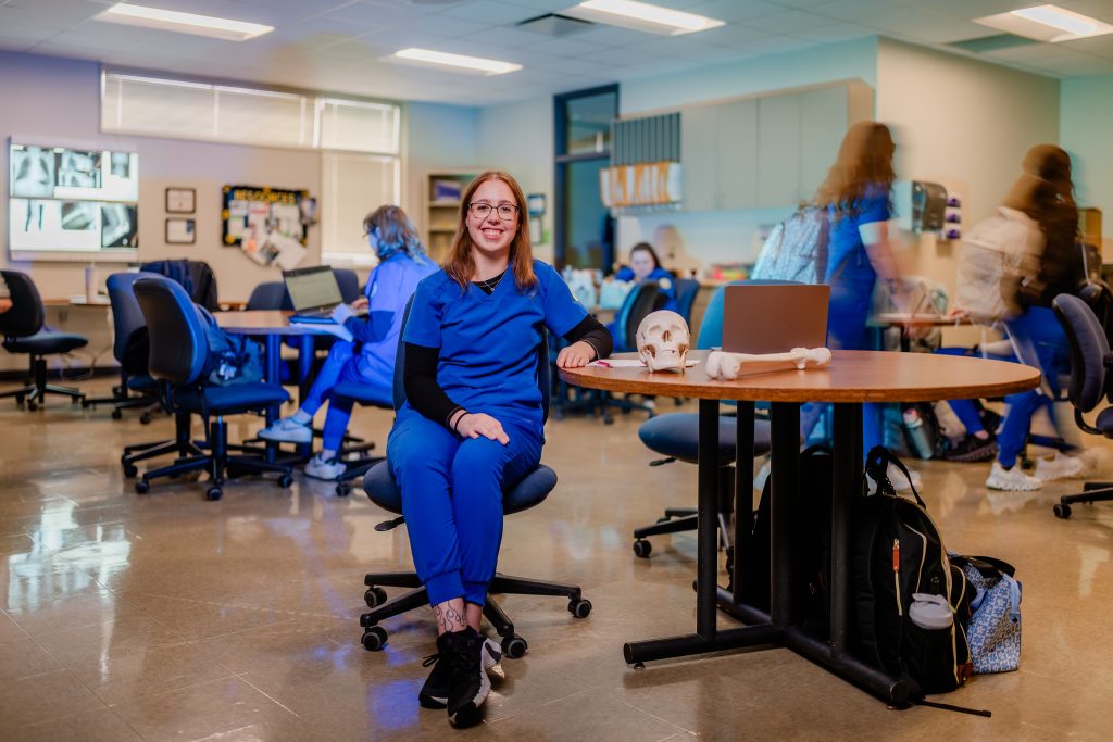 Happy nursing student in scrubs sitting at a table with a laptop and skeleton on it.