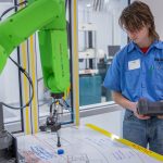 High school student controlling a green robotic arm with a remote control.