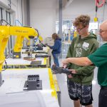 High school student controlling a yellow robotic arm with a remote control.