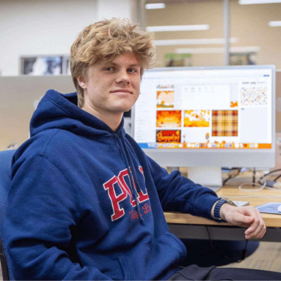 Graphic design student Adam is happily sitting at his work station with several computer screens.