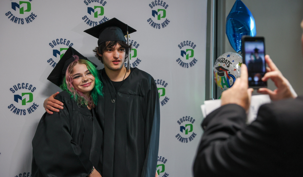Journey student couple stands in front of MPTC backdrop in their cap and gowns.