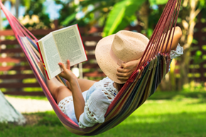 Person wearing a hat laying in a hammock reading a book.