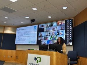 Presenter, Leslie Laster, stands at a podium, presenting to K-12 educators, with a screen of virtual attendees behind her. 