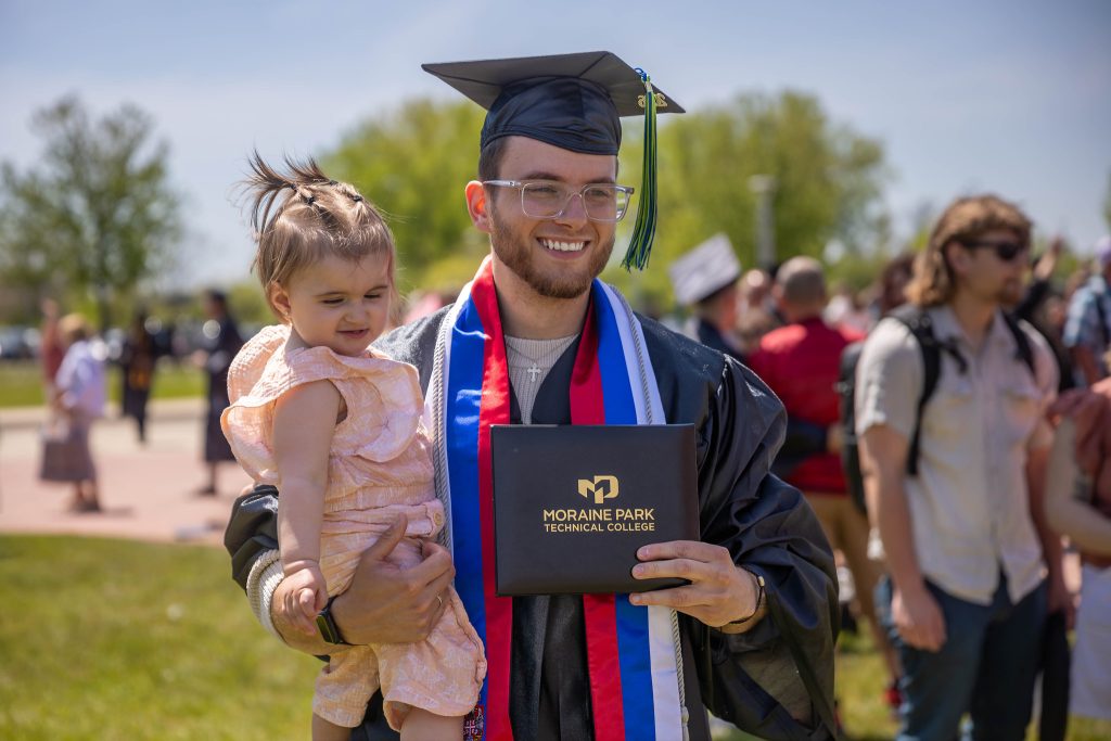 Smiling student in graduation gown holding his degree in one hand and a child in the other hand.