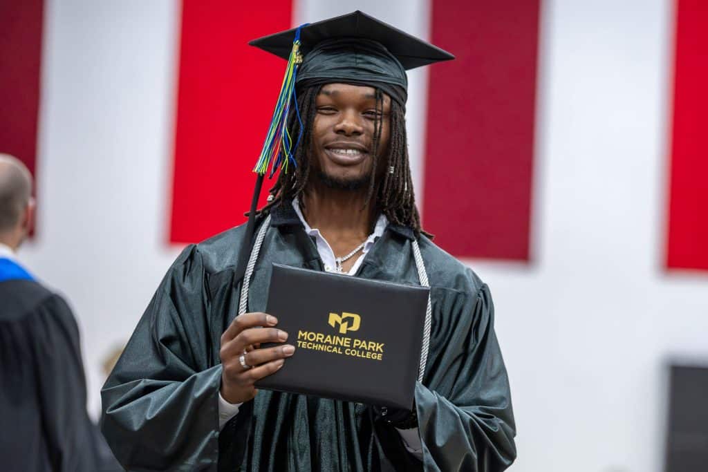 Smiling student showing their Moraine Park diploma wearing their graduation gown.