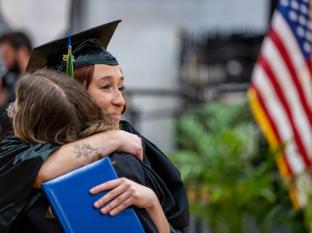 Graduates hugging at commencement in front of American flag.
